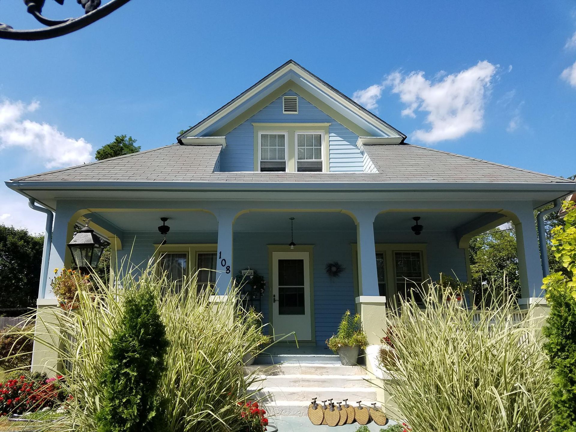 Blue cottage with a front porch, white trim, and tall overgrown plants in the yard under a bright sky