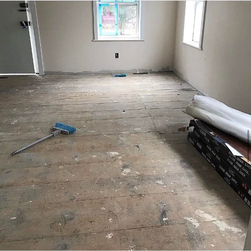 Empty room with unfinished wooden floor, windows, and construction materials stacked along the right wall.