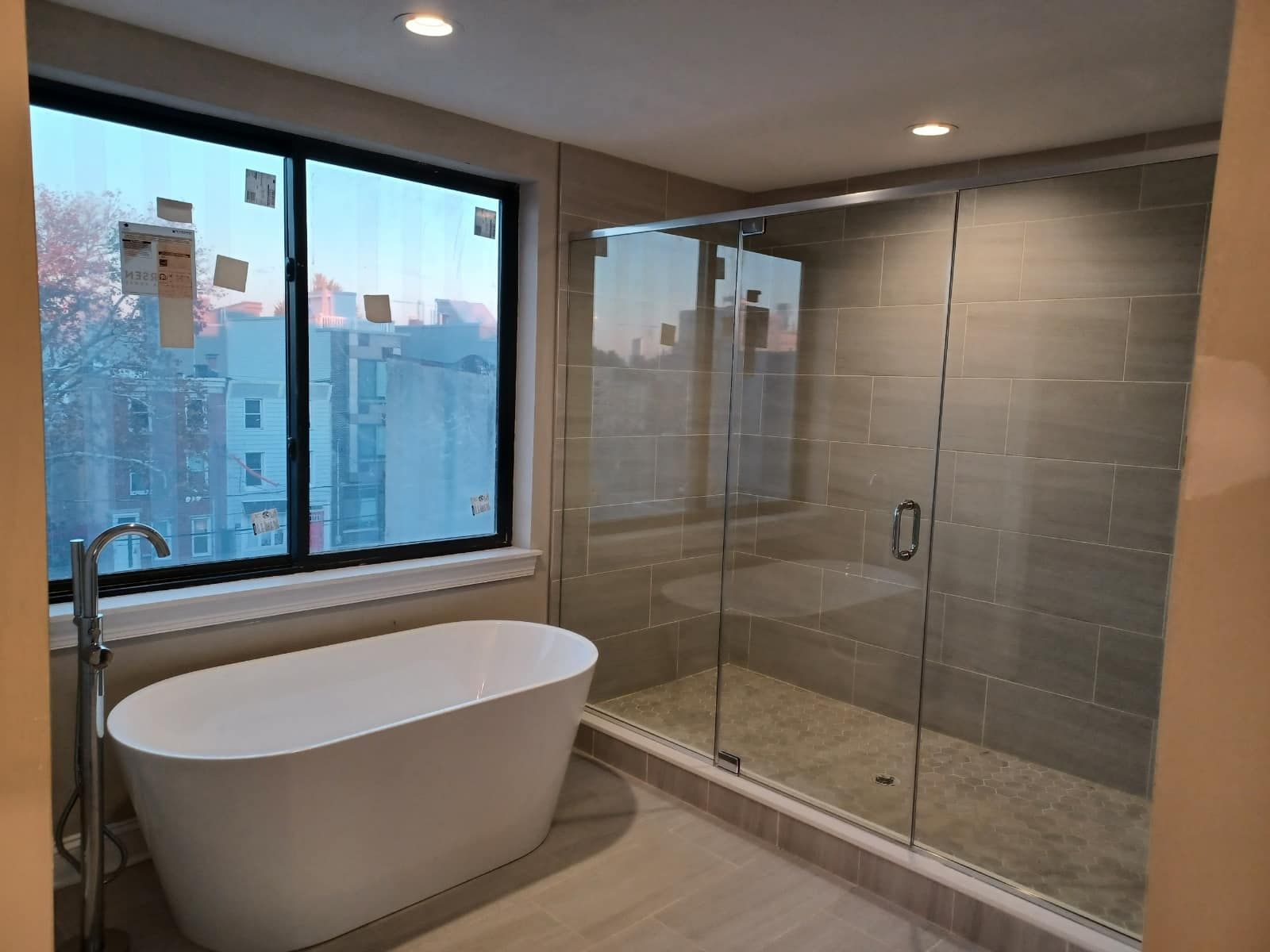 Modern bathroom with freestanding tub, glass shower, and city view through a large window