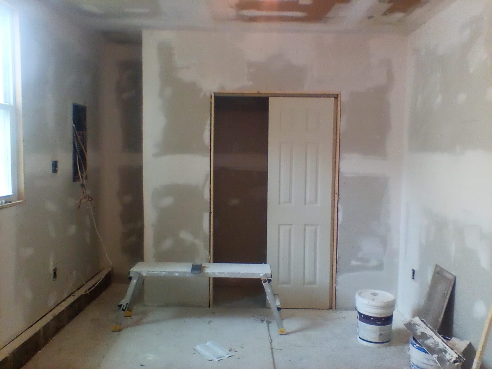 Empty room under renovation with drywall patches, a window, doorway, sawhorse, and paint bucket.