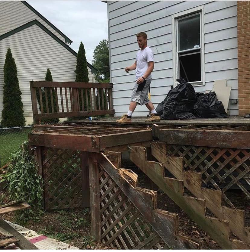 Man standing on a damaged wooden deck beside a house, with debris and black trash bags nearby.