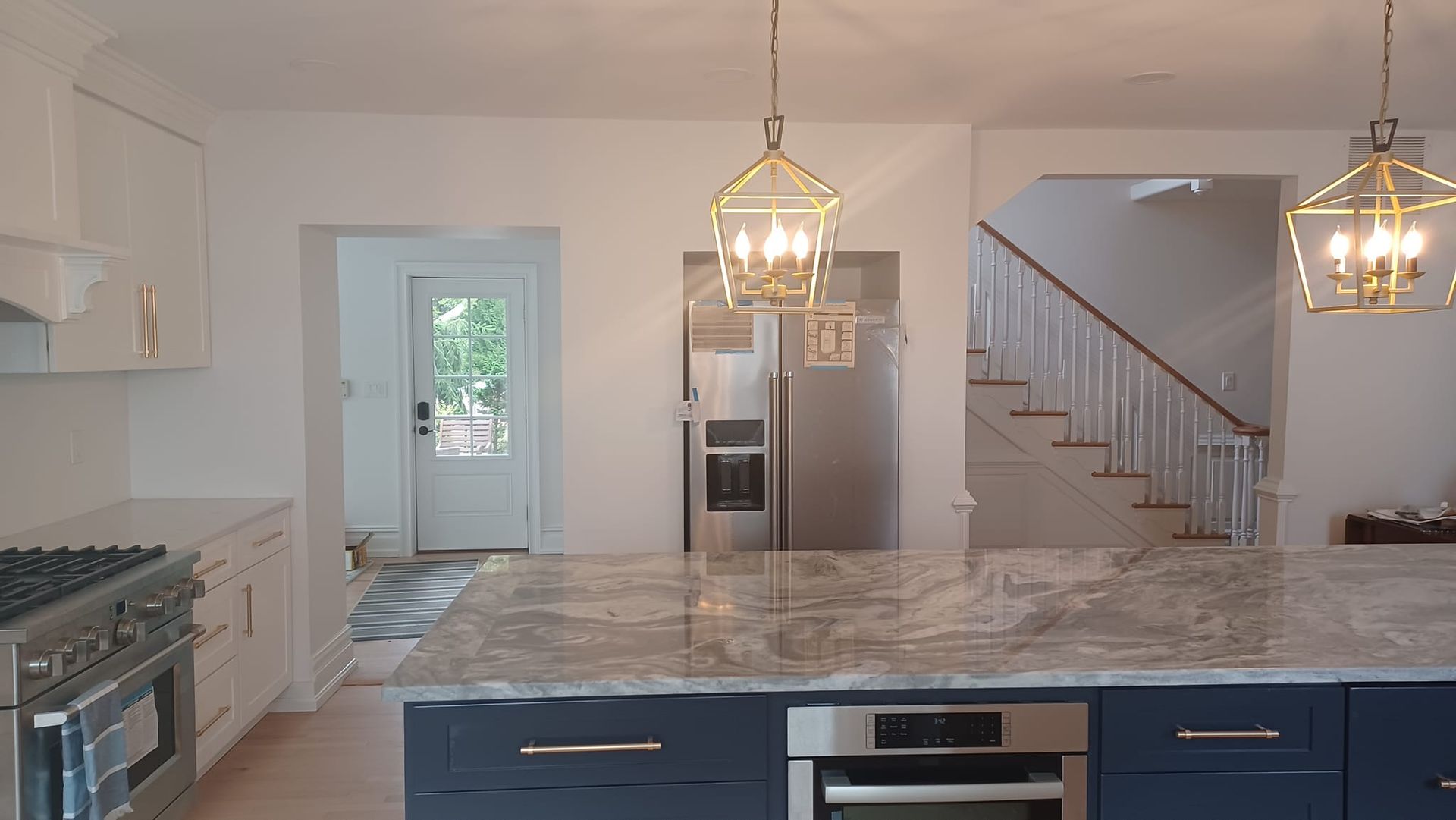 Modern white kitchen with blue island, marble countertops, pendant lights, and staircase in background