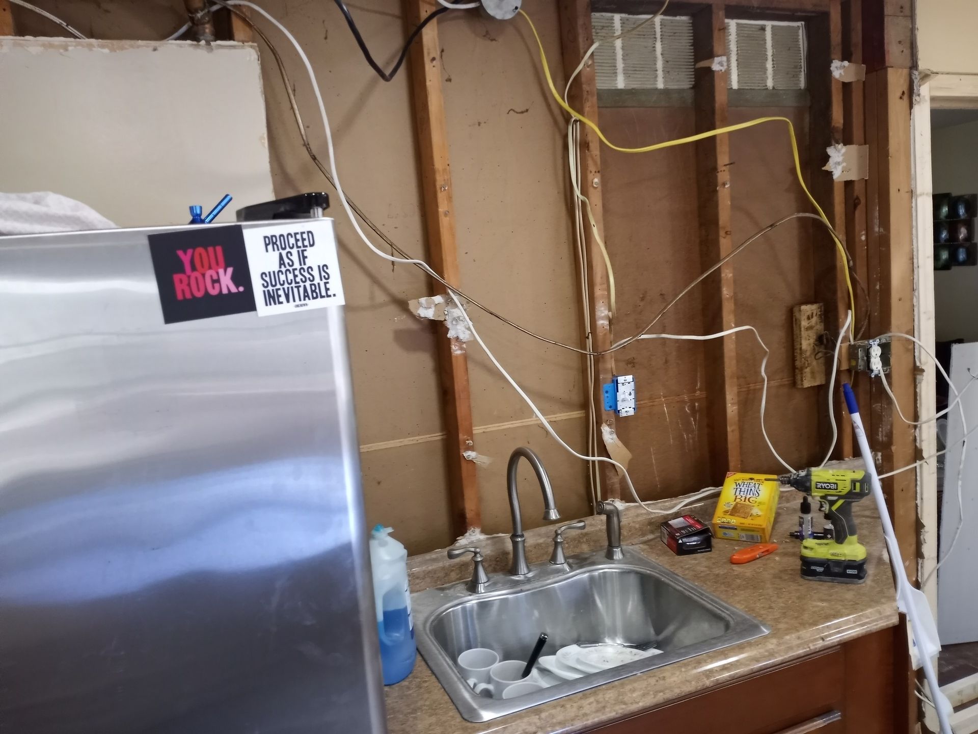 Kitchen sink and refrigerator against exposed wall studs, with hanging wires and tools during renovation.