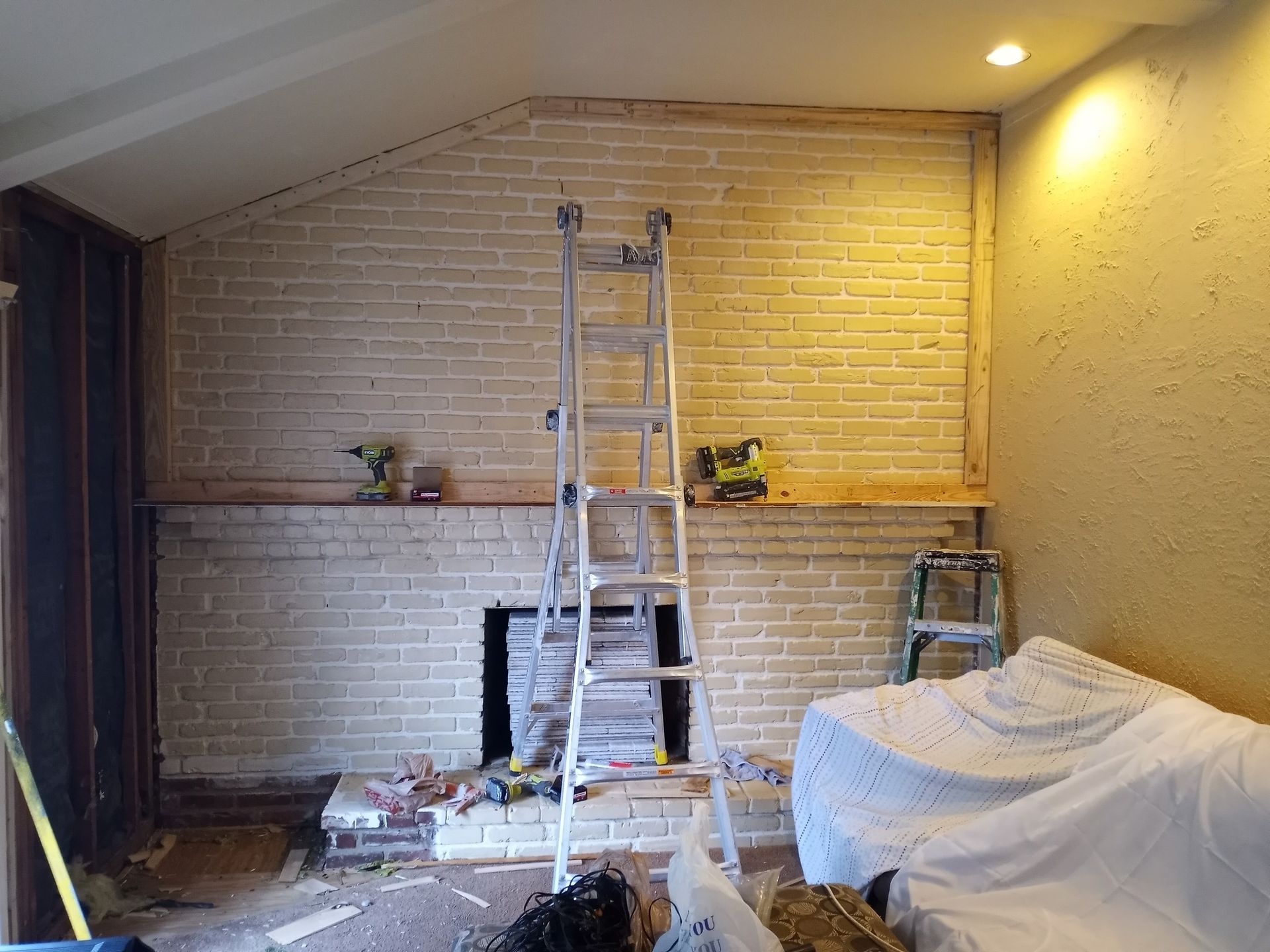 Living room with a brick fireplace, ladder, and furniture covered in protective sheets during renovation