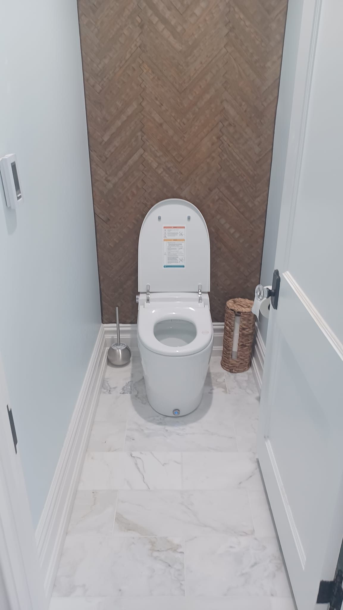 Small bathroom with a toilet, wood accent wall, and white marble-look floor and walls.