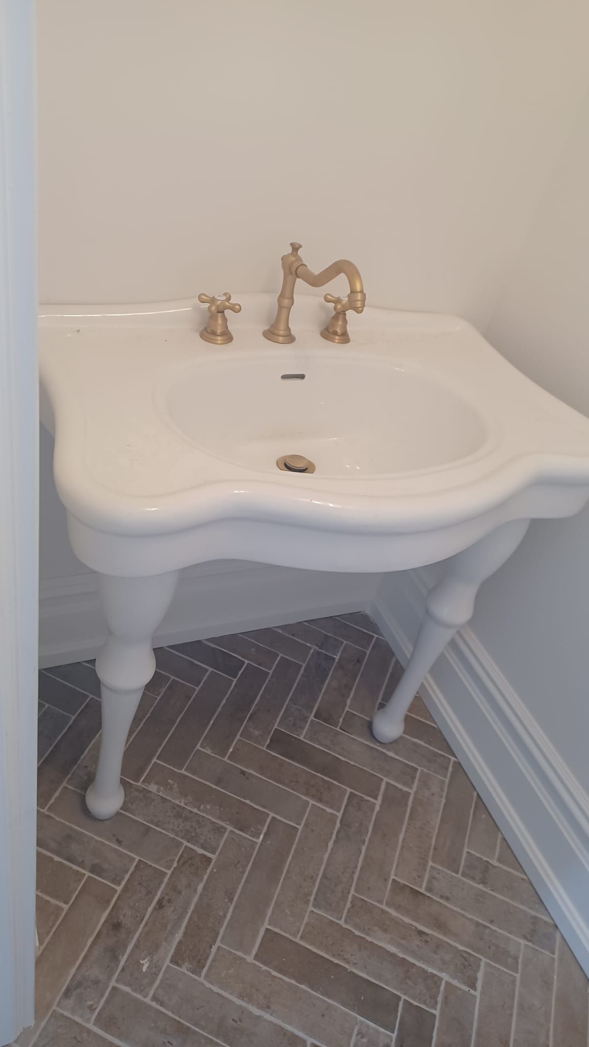 White pedestal sink with brass faucet in a small bathroom corner over herringbone tile flooring.