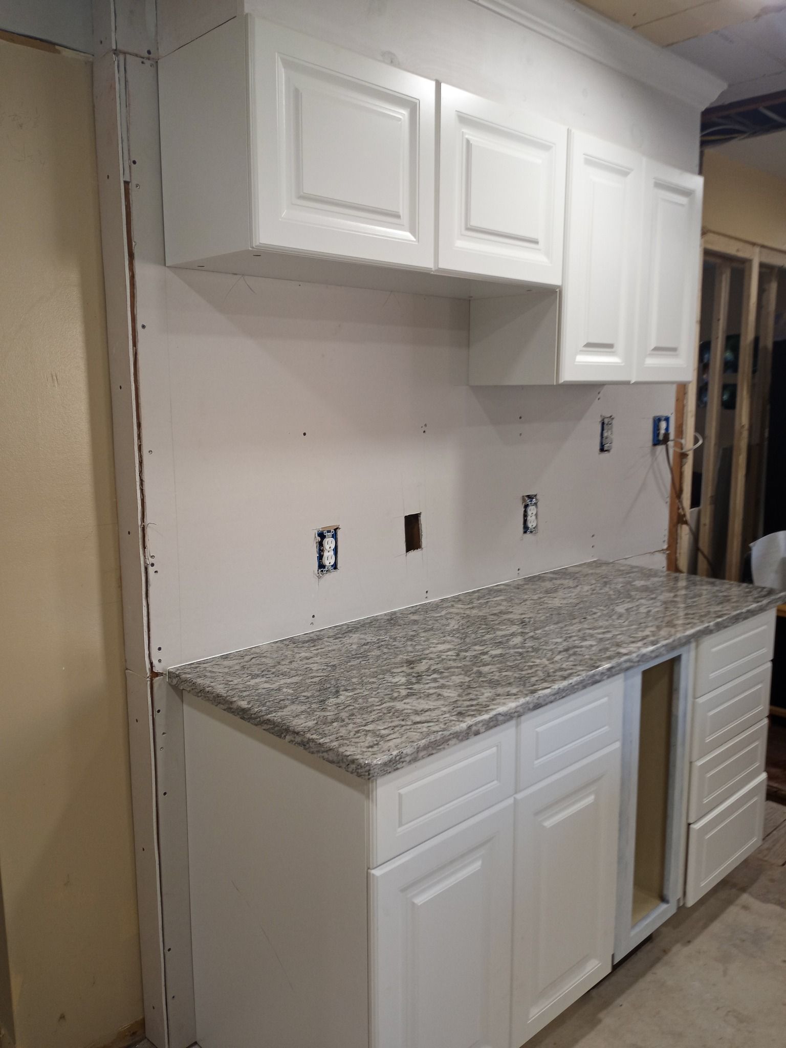 Unfinished kitchen wall with white cabinets, granite countertop, and lower cabinets installed along one side.