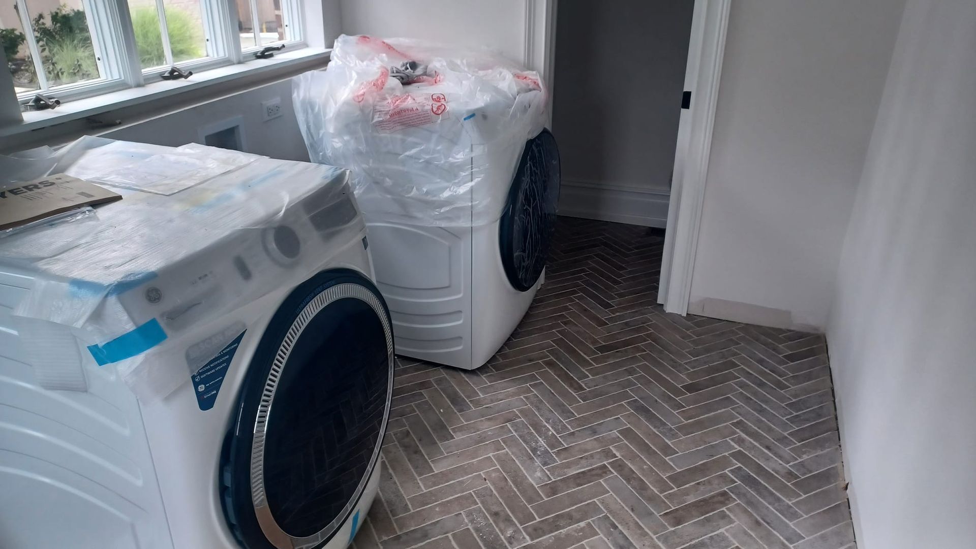 Two front-loading washers in a narrow laundry room, one partly wrapped in plastic.