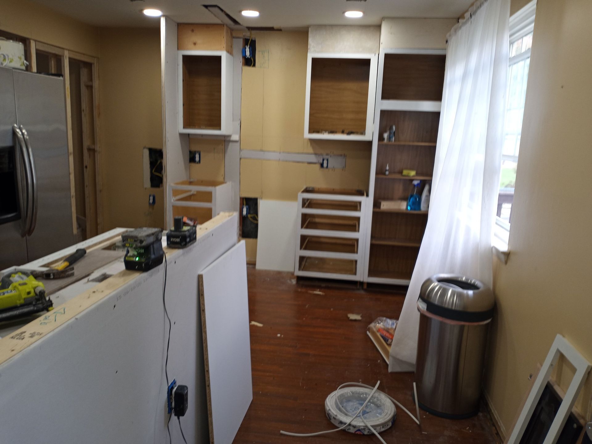 Empty kitchen under renovation with white cabinets, wood floor, and a trash can by the window