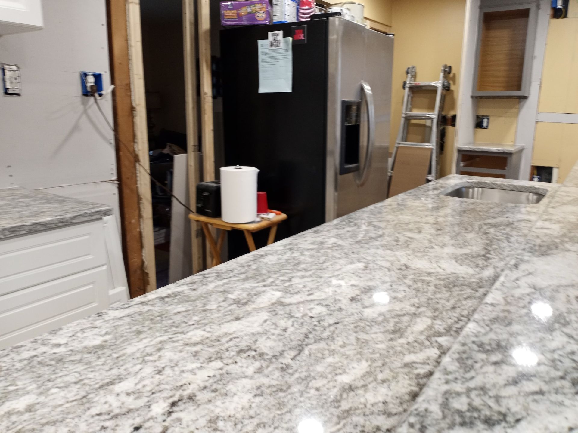 Kitchen counter with granite top, refrigerator, paper towels, and step ladder in a partly unfinished room