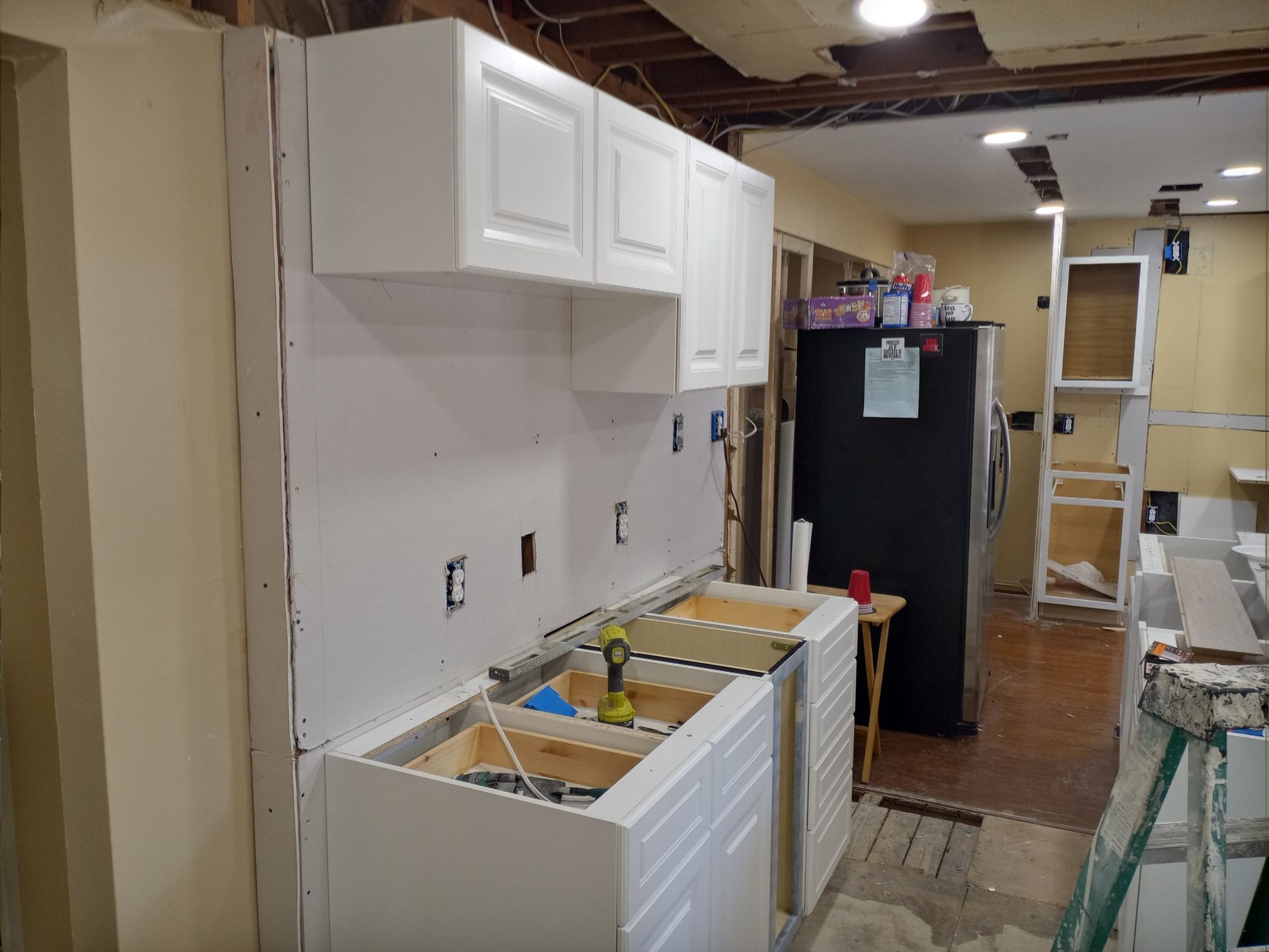 Kitchen under renovation with white cabinets, exposed wiring, and a black refrigerator in the background