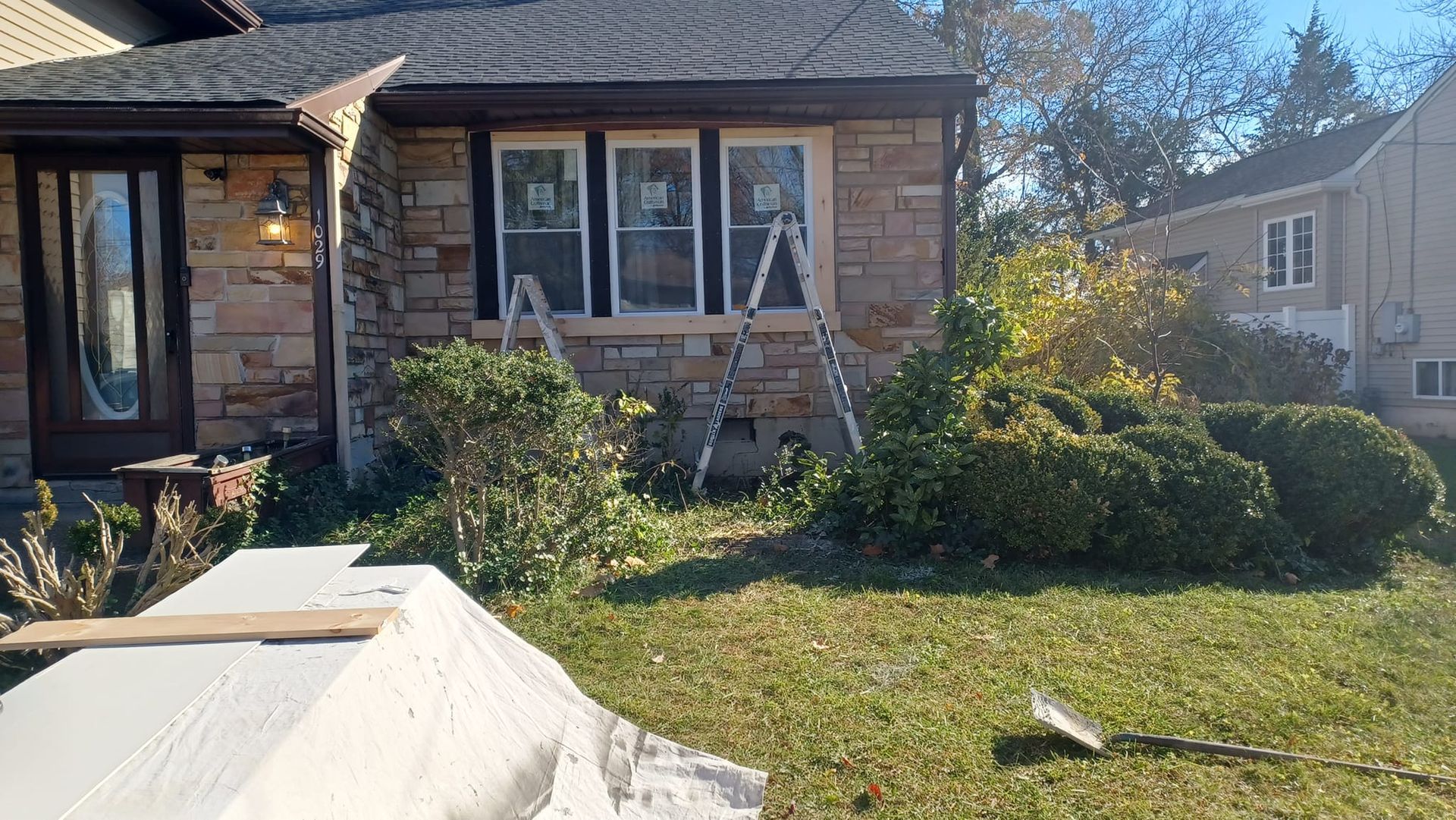 Single-story stone house with front windows, overgrown yard, and a white-covered object in the foreground.