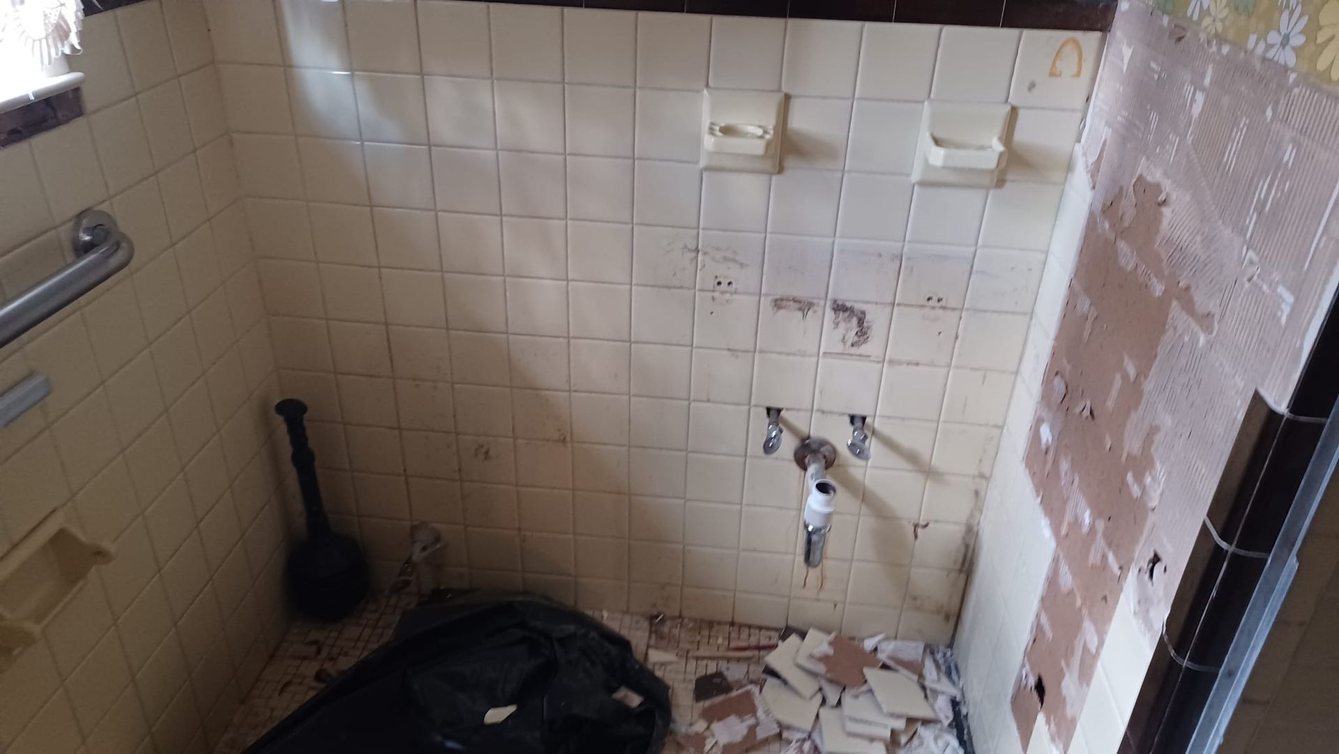 Small tiled bathroom with a damaged wall, exposed pipes, and debris on the floor.