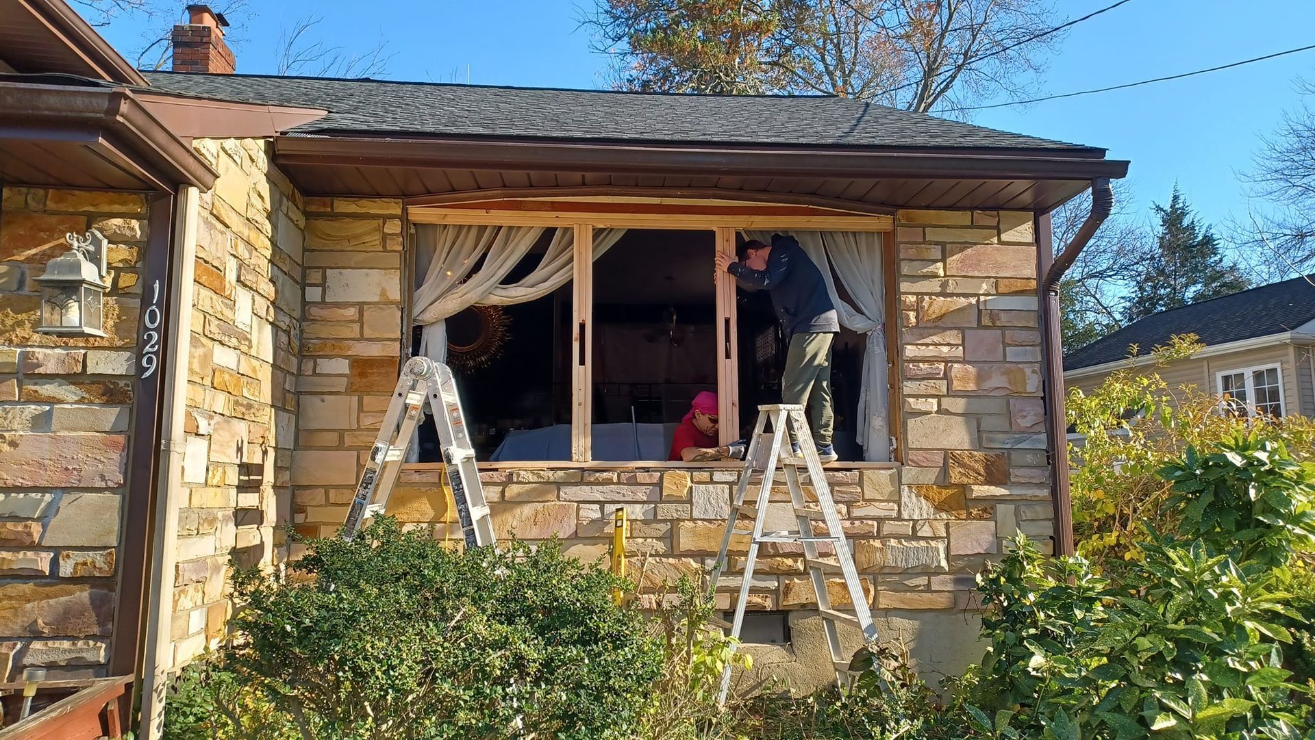 Two ladders under a stone house window during home renovation, with curtains and exposed interior visible.