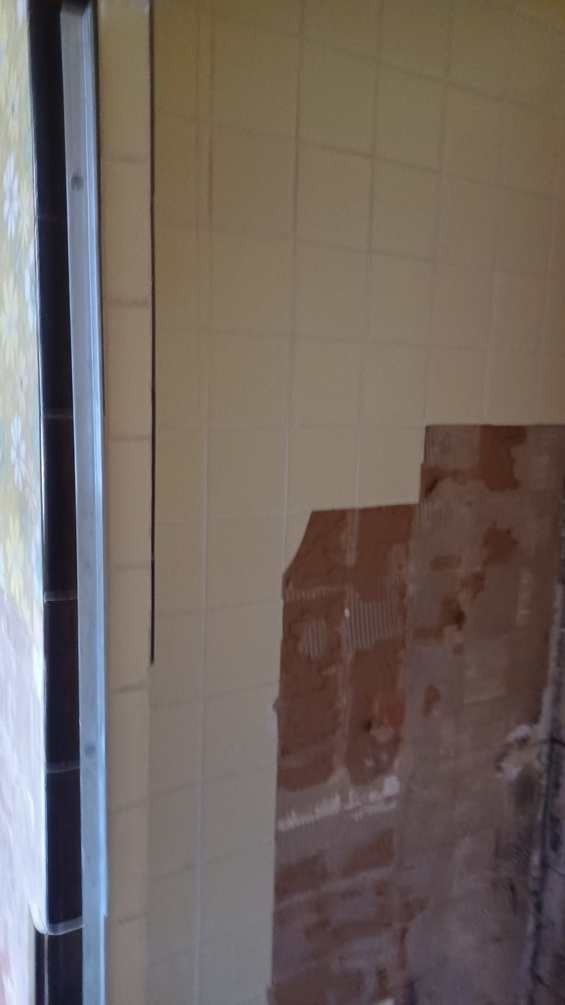 Partially stripped beige tiled wall with exposed red brick near a doorway