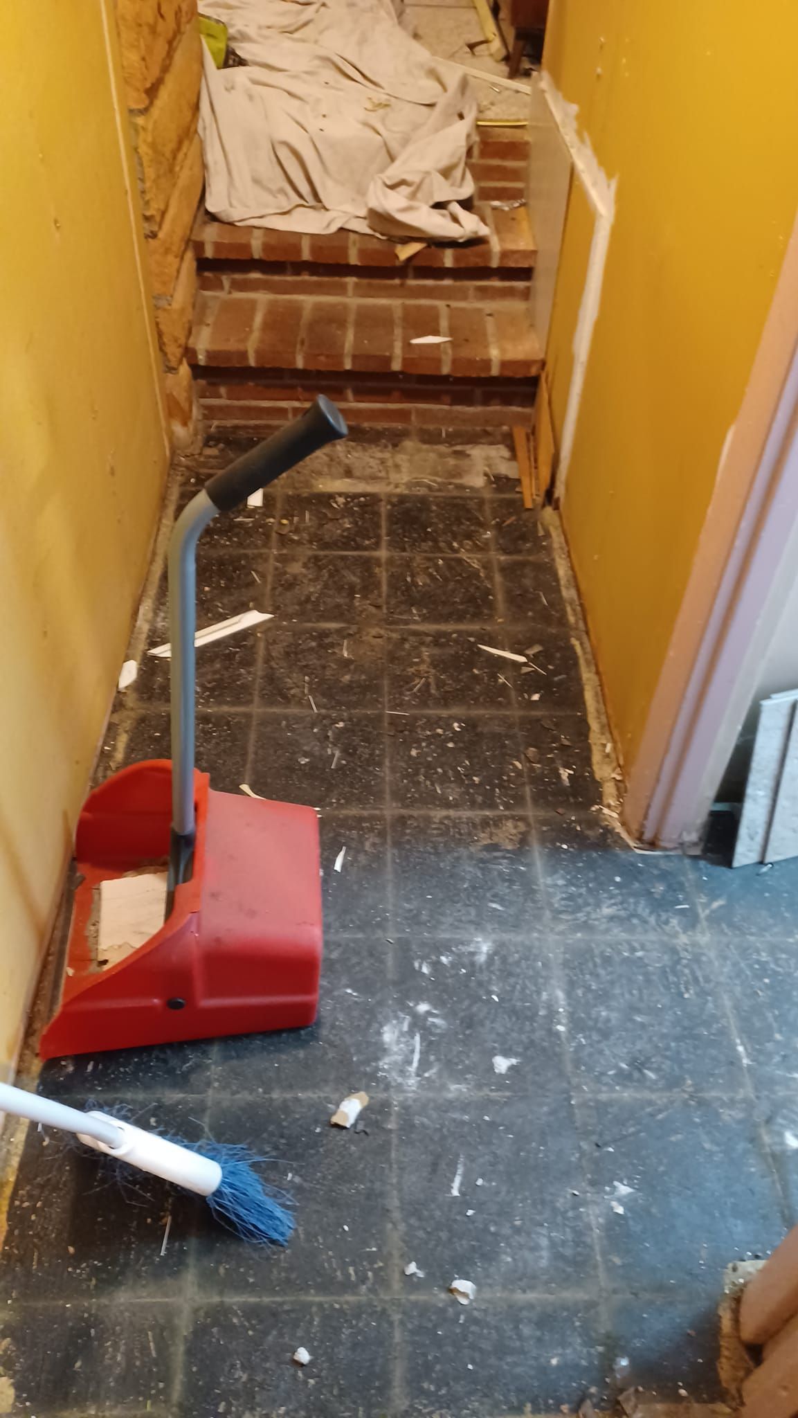 Narrow hallway with construction debris, a red dustpan, and a vacuum hose on a damaged floor.