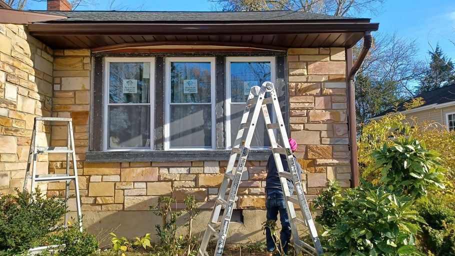Ladder propped against a stone house exterior beneath a window, with bushes in front.