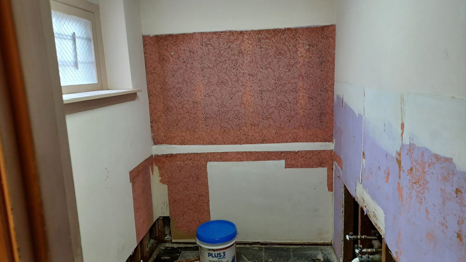 Small bathroom under renovation with exposed wall patching, a window, and a blue bucket on the floor