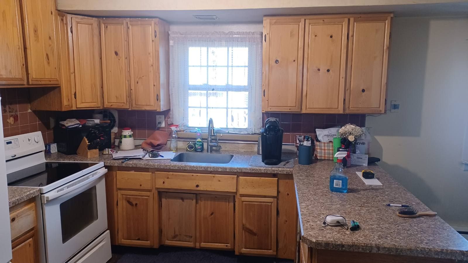 Wooden kitchen with white stove, center sink under window, and granite countertop cluttered with items.