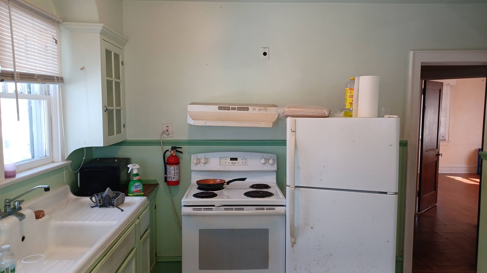 Small kitchen with white stove and refrigerator, light green walls, sink by window, and doorway to another room.