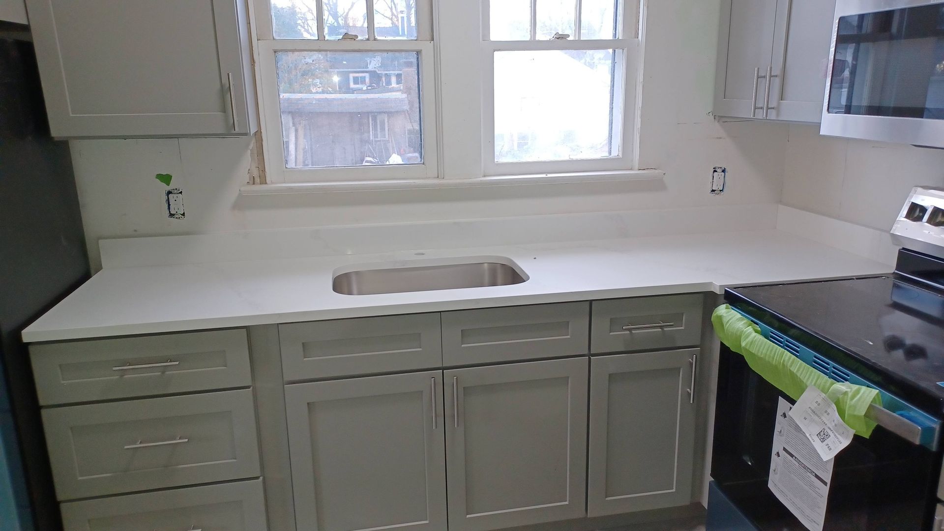 Kitchen with white countertop, gray cabinets, sink, and window above, next to a stove with a green towel