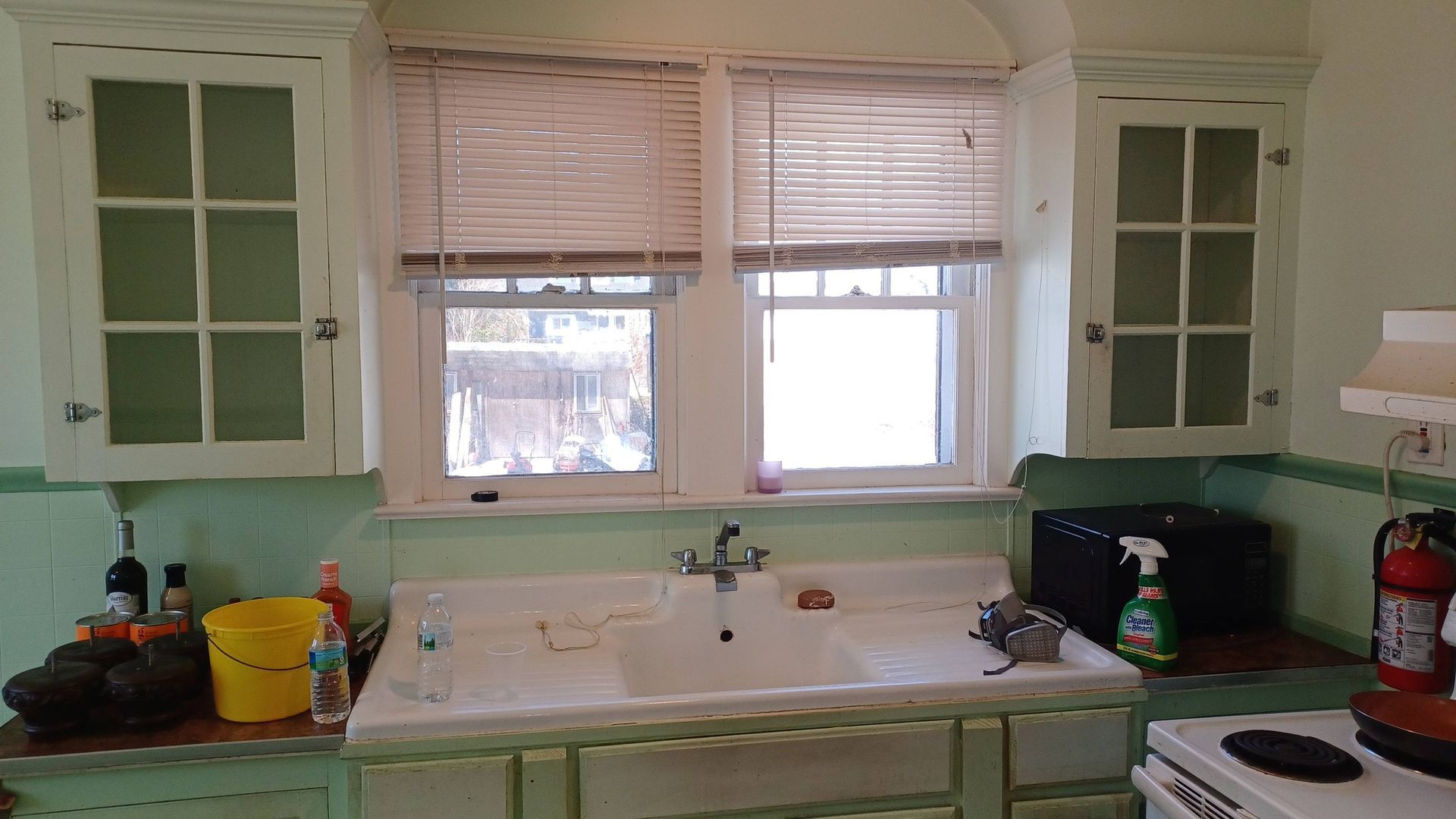 Kitchen sink beneath windows, with pale green cabinets and countertops cluttered with dishes and cleaning items.