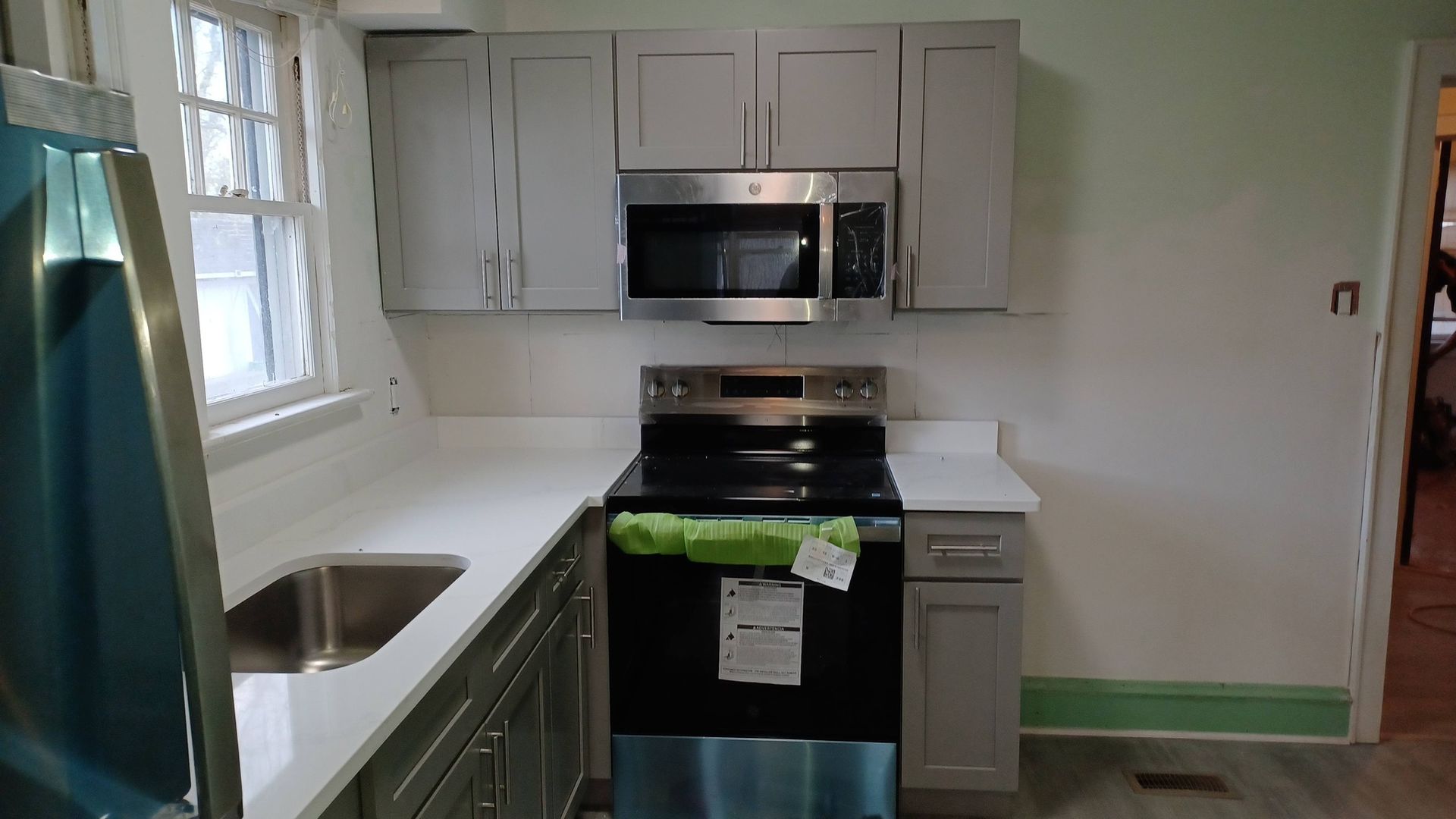 Small kitchen with white cabinets, stainless sink and microwave, black stove, and green painter’s tape on the baseboards