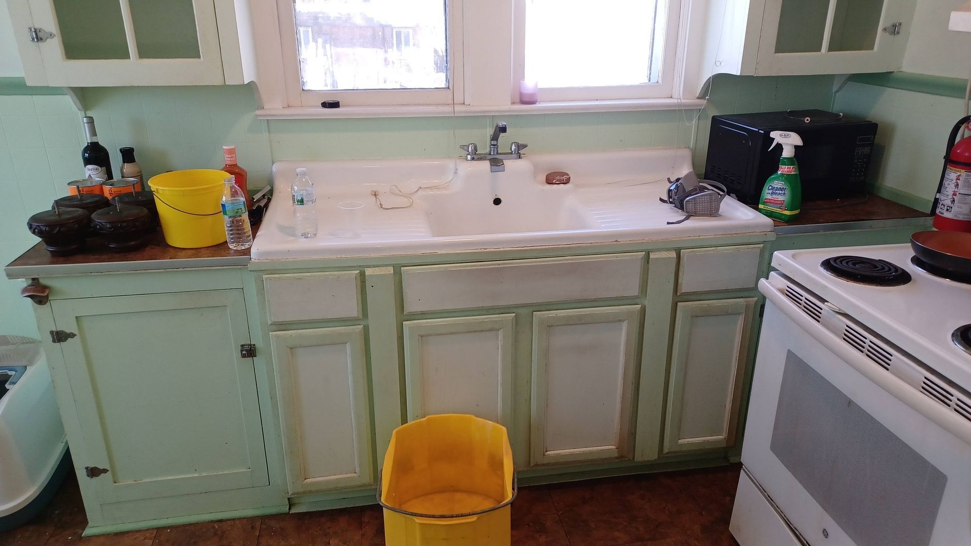 Small kitchen with mint cabinets, white sink under two windows, and a yellow trash bin in front.