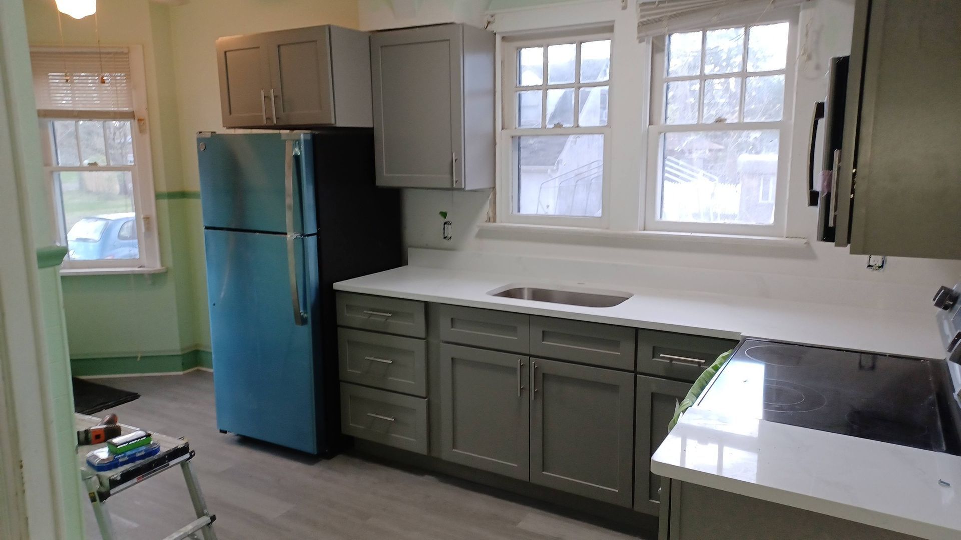 Bright kitchen with gray cabinets, blue refrigerator, white countertops, and large windows.