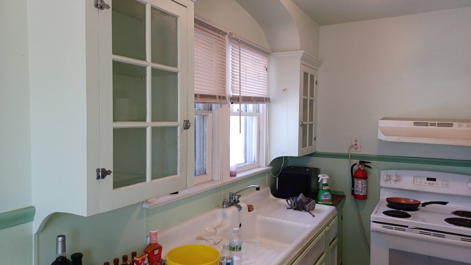 Small kitchen with mint-green cabinets, sink, window blinds, and a white stove with a red fire extinguisher nearby