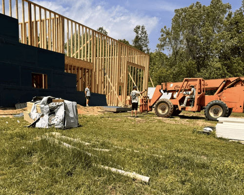 Construction site with a wooden building frame, orange loader, and materials on grass under a clear sky