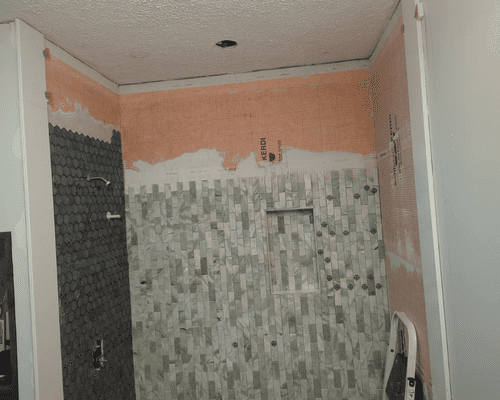 Partially tiled bathroom wall under renovation, with pink upper walls and gray mosaic tiles below.