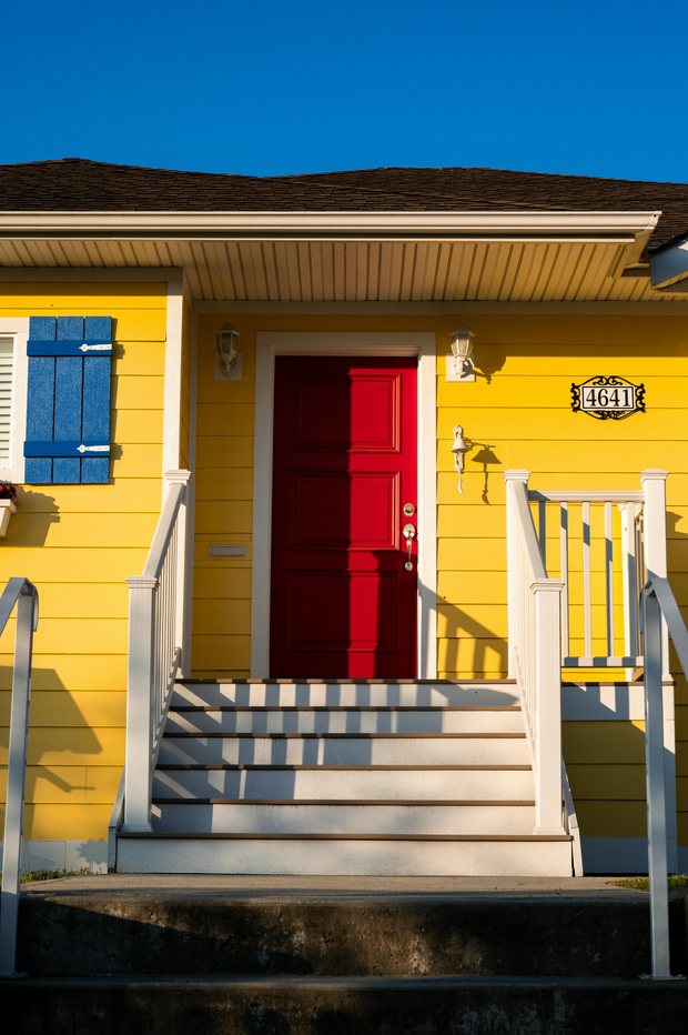 Yellow house porch with red door, blue shutters, and white railing under a sunny roofline