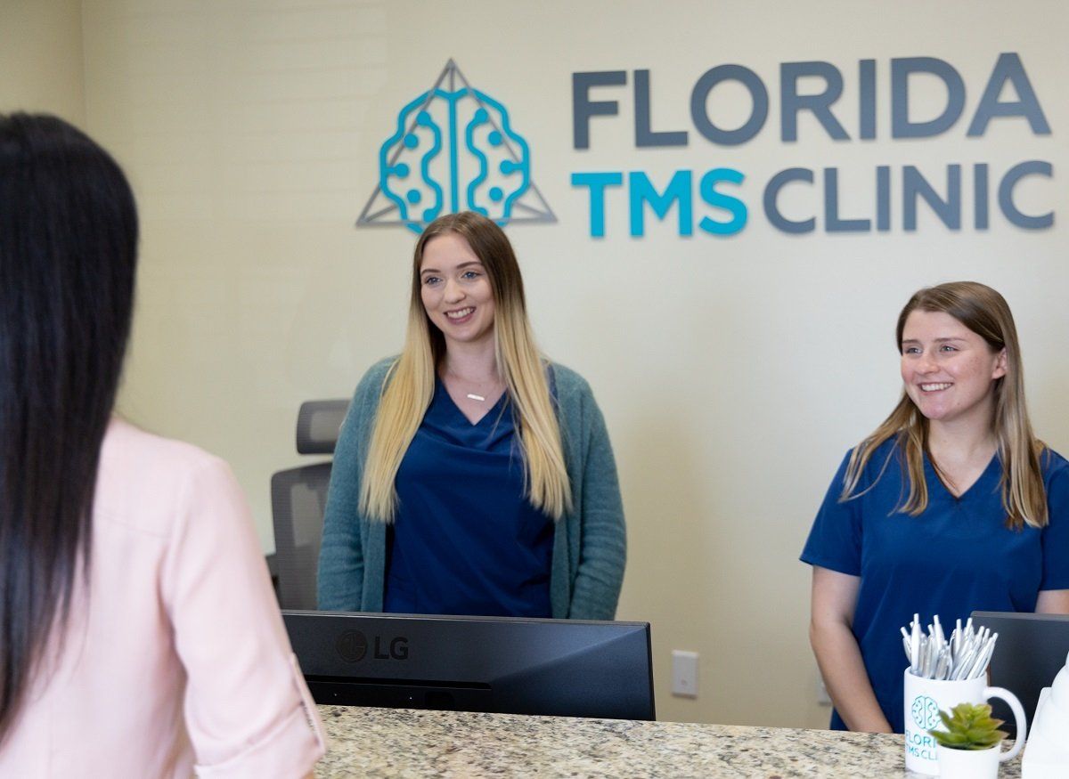 Two smiling receptionists at the Florida TMS Clinic desk greet a patient. The clinic's logo and name are visible on the wall behind them.
