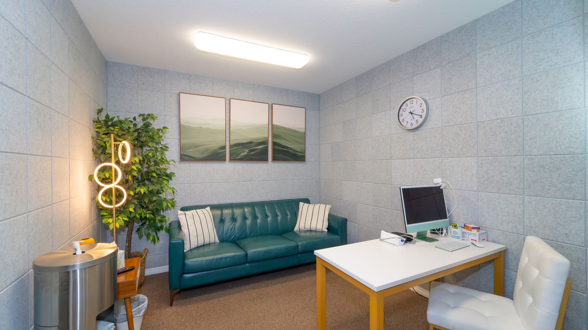 A cozy office space with a teal couch, desk, and artwork. A plant and a clock are visible, along with gray textured walls.