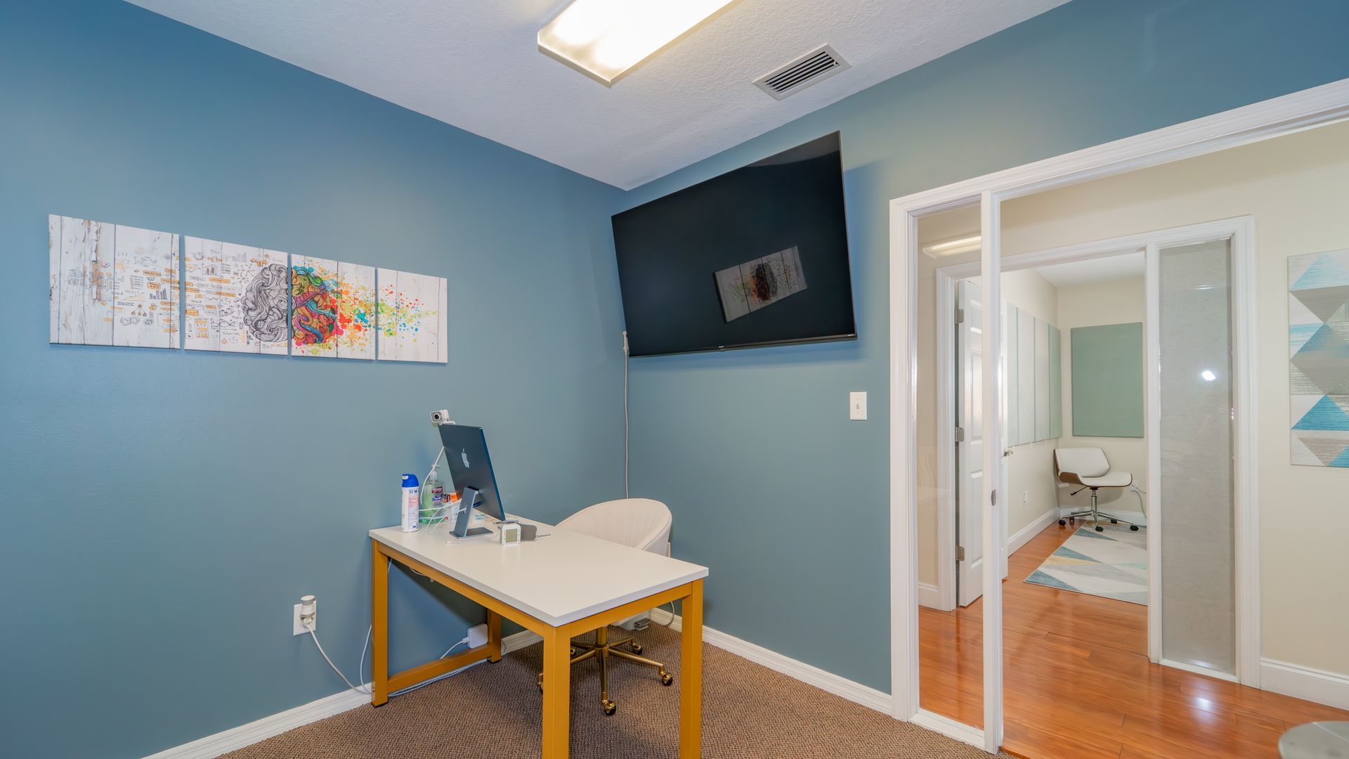 A doctor's office with blue walls, a desk, computer, TV, and a partial view of another room with a patient chair.