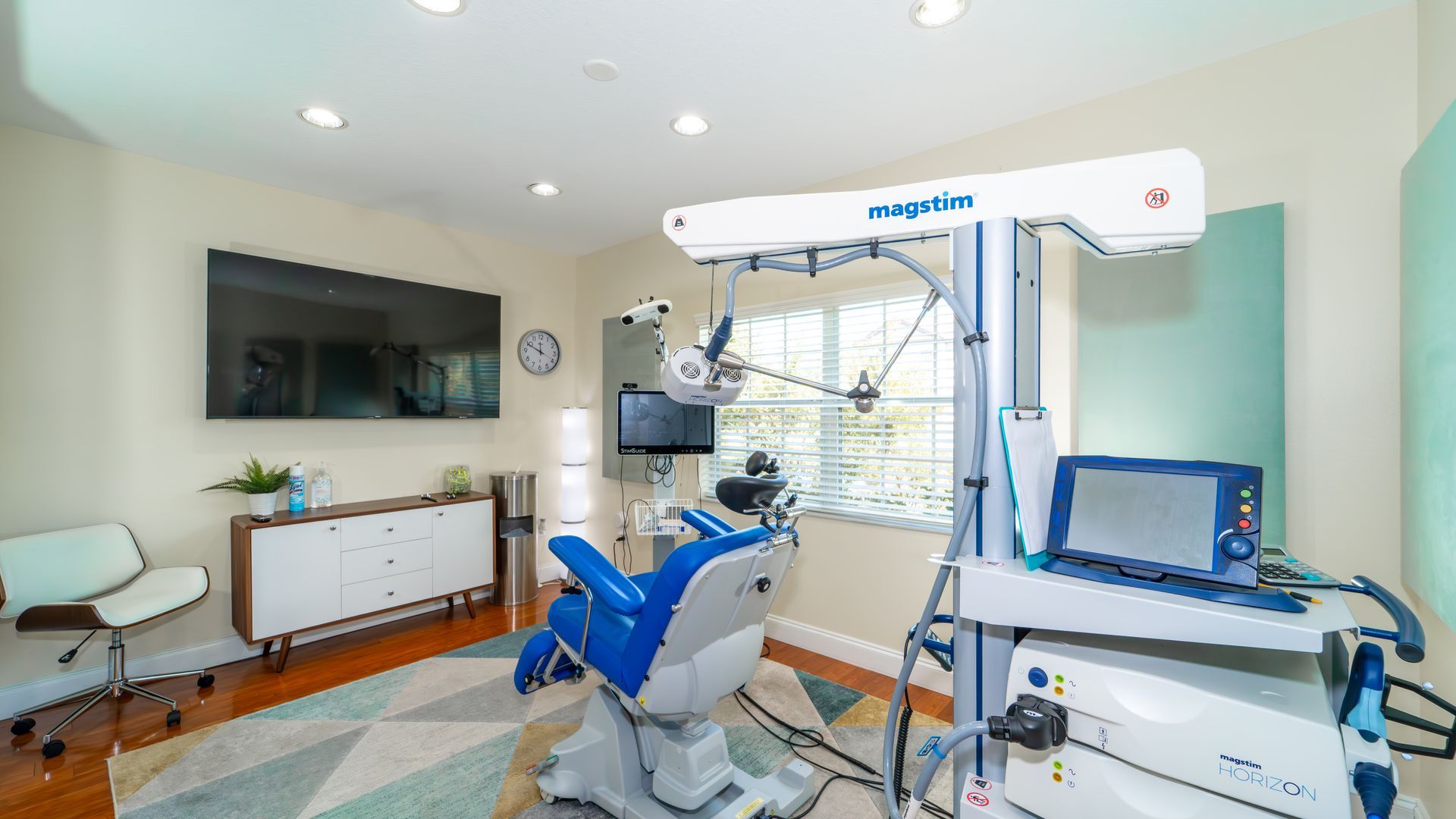 A medical room with blue equipment, a patient chair, and a machine with a screen. Light colors with a mid-century modern accent.
