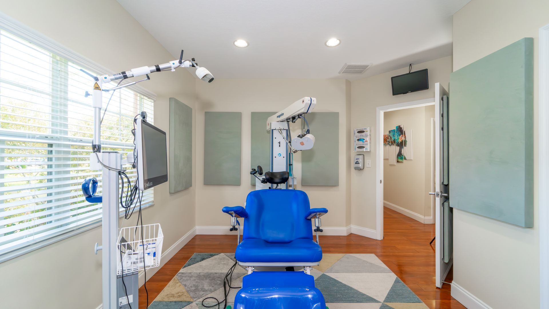 Examination room with a blue medical chair, diagnostic equipment, and a window with blinds. The room has wooden floors and beige walls.