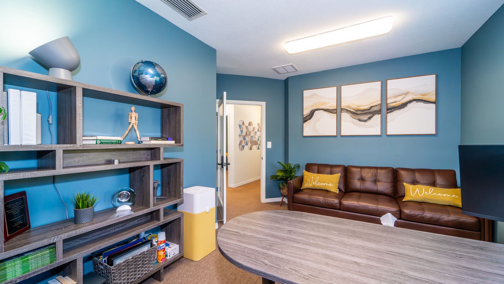 An office with blue walls, a brown leather sofa, and a round desk. There's a shelving unit with decor and a door leading to another area.