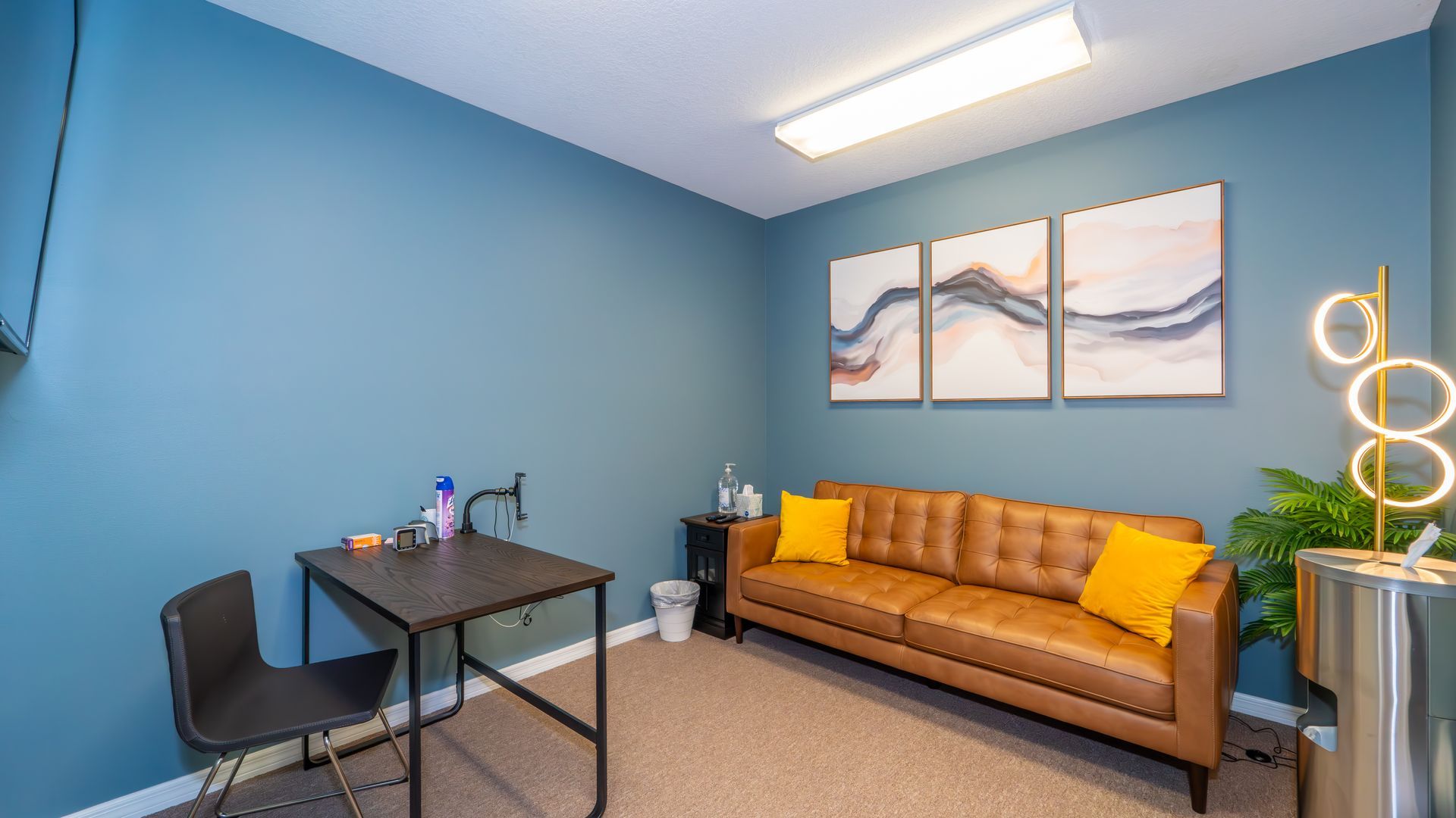 A small office with blue walls, a brown leather couch with yellow pillows, and a small desk. Artwork hangs above the couch.