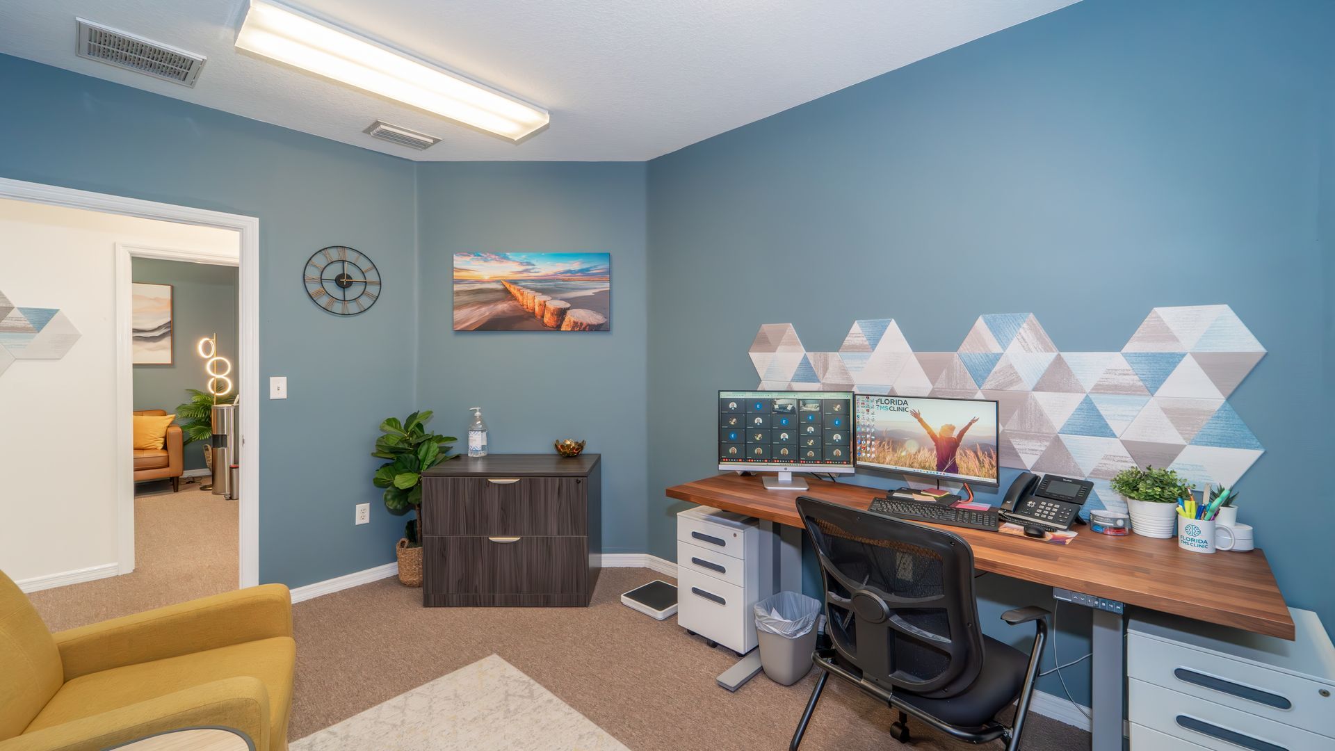 An office space with blue walls, a desk with two monitors, and a yellow armchair. Another room is visible in the background.