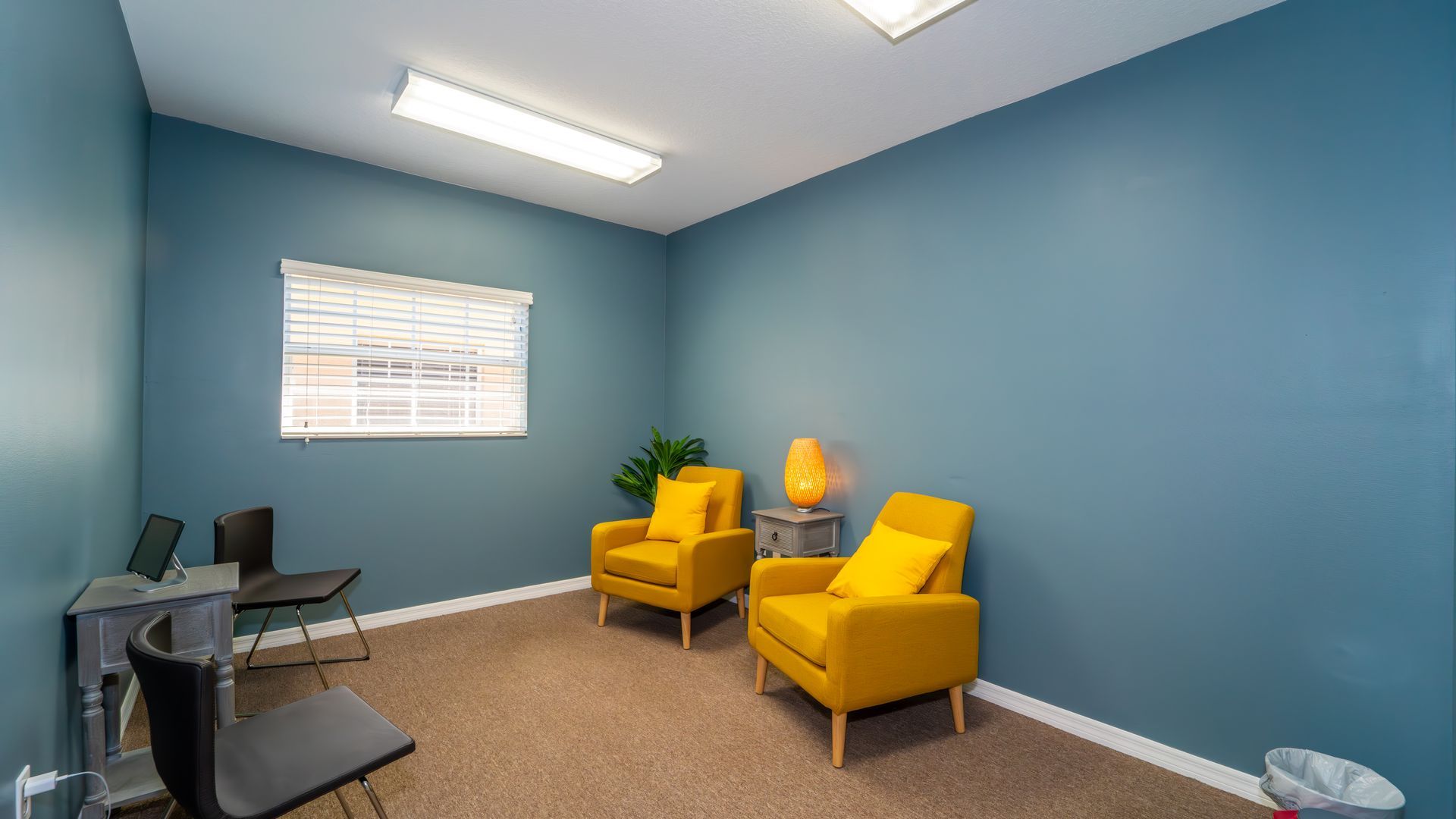 A waiting room with two yellow armchairs, a small table with a lamp, and a desk with chairs, all against blue walls.
