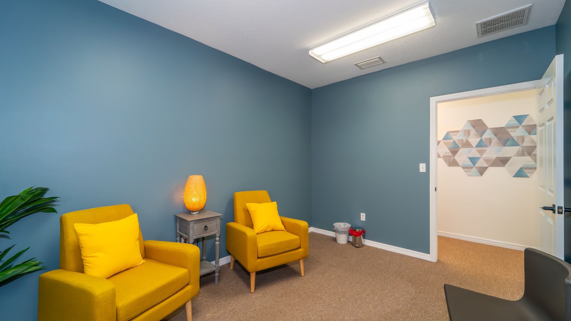 A counseling office with two yellow armchairs, a side table, and a door with a geometric pattern. Blue walls and tan carpet.