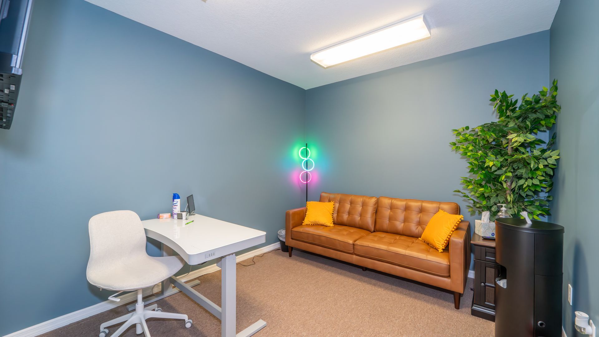 A modern office room with a blue wall, a brown leather sofa, a white desk, and an accent light display.