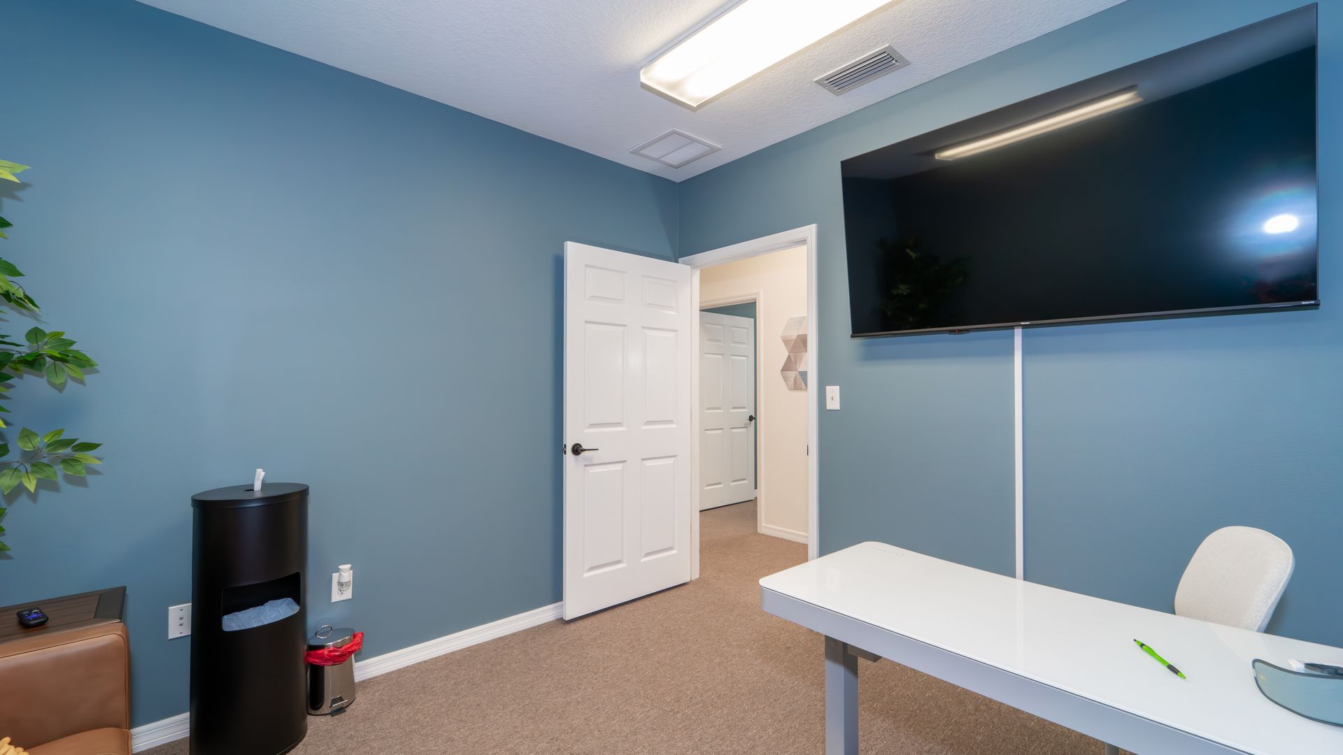 Office interior with blue walls, a large TV, a white desk, and a trash can. A door leads to another room.