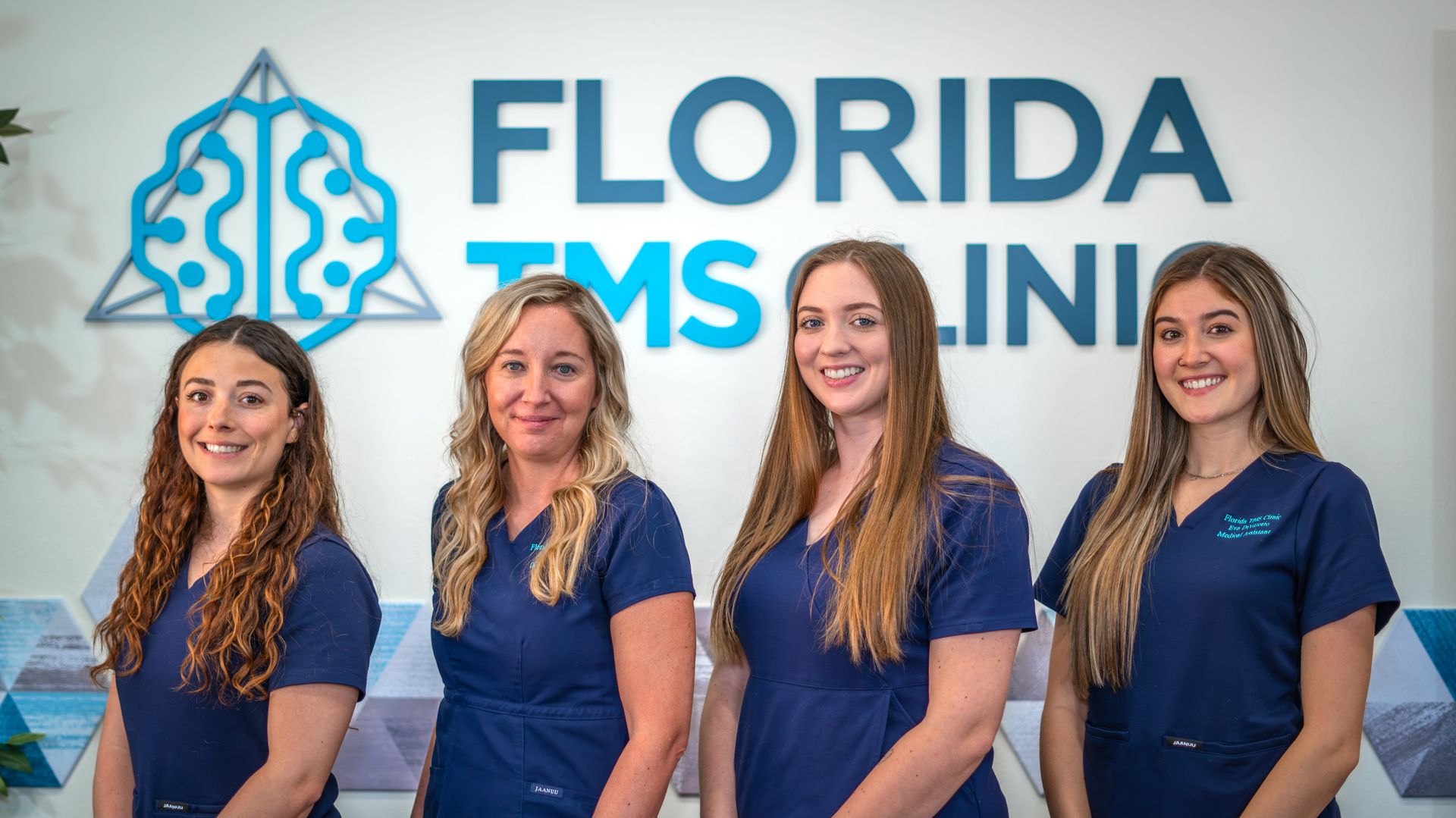 Four women in blue scrubs stand in front of a 