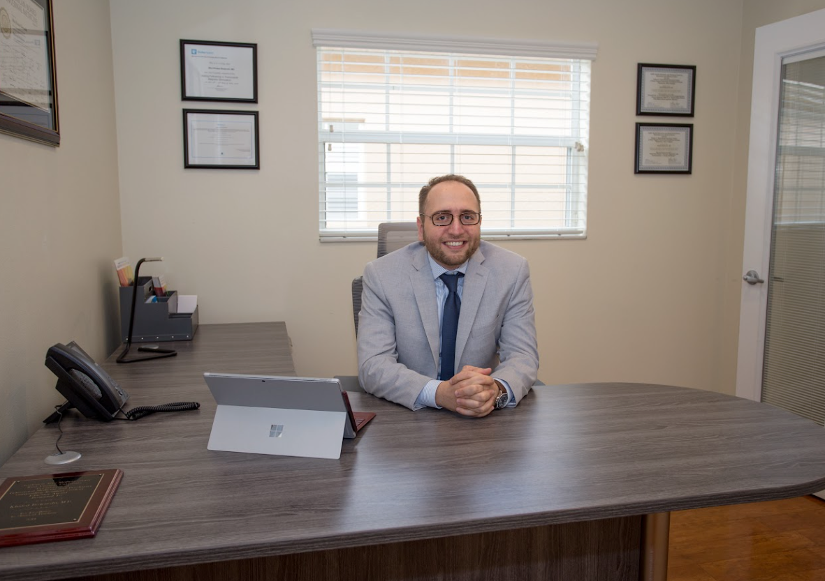 Man seated behind a large wooden desk in an office, wearing a light blue shirt and tie.
