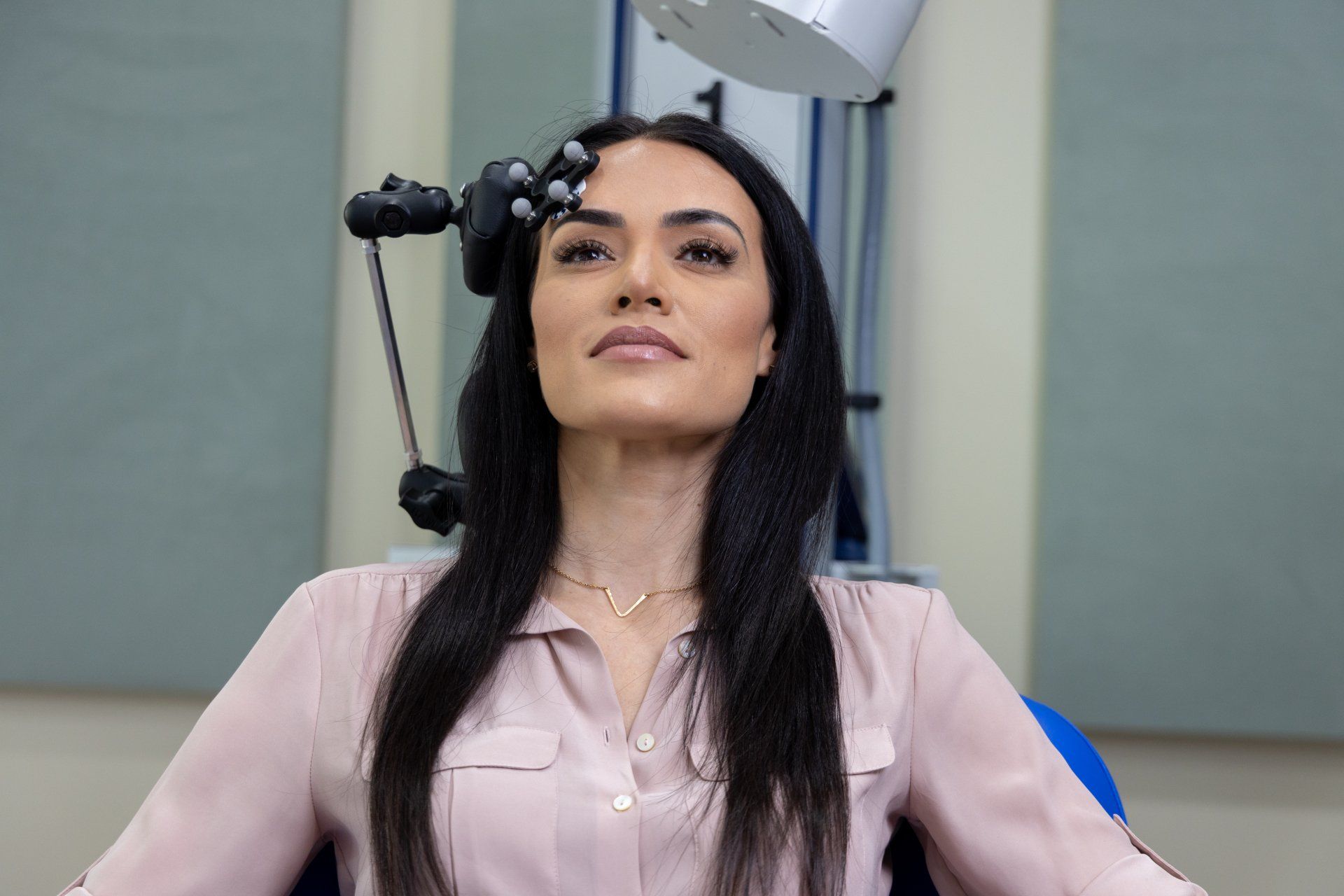 A woman sits with a device above her head, looking upwards. She has dark hair and a pink shirt in a clinical setting.