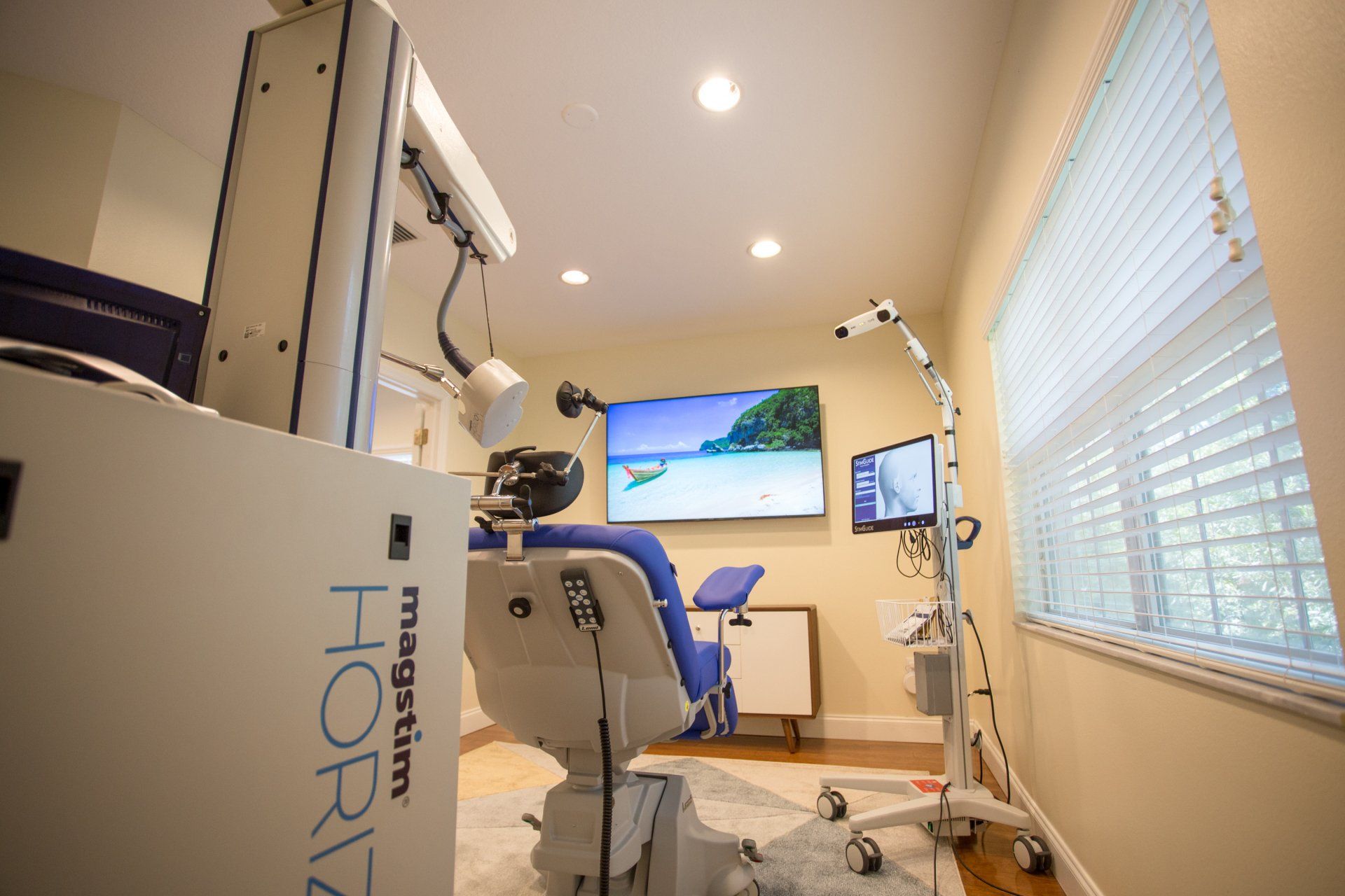 A room with a medical chair and equipment for transcranial magnetic stimulation (TMS). A screen shows a beach scene.