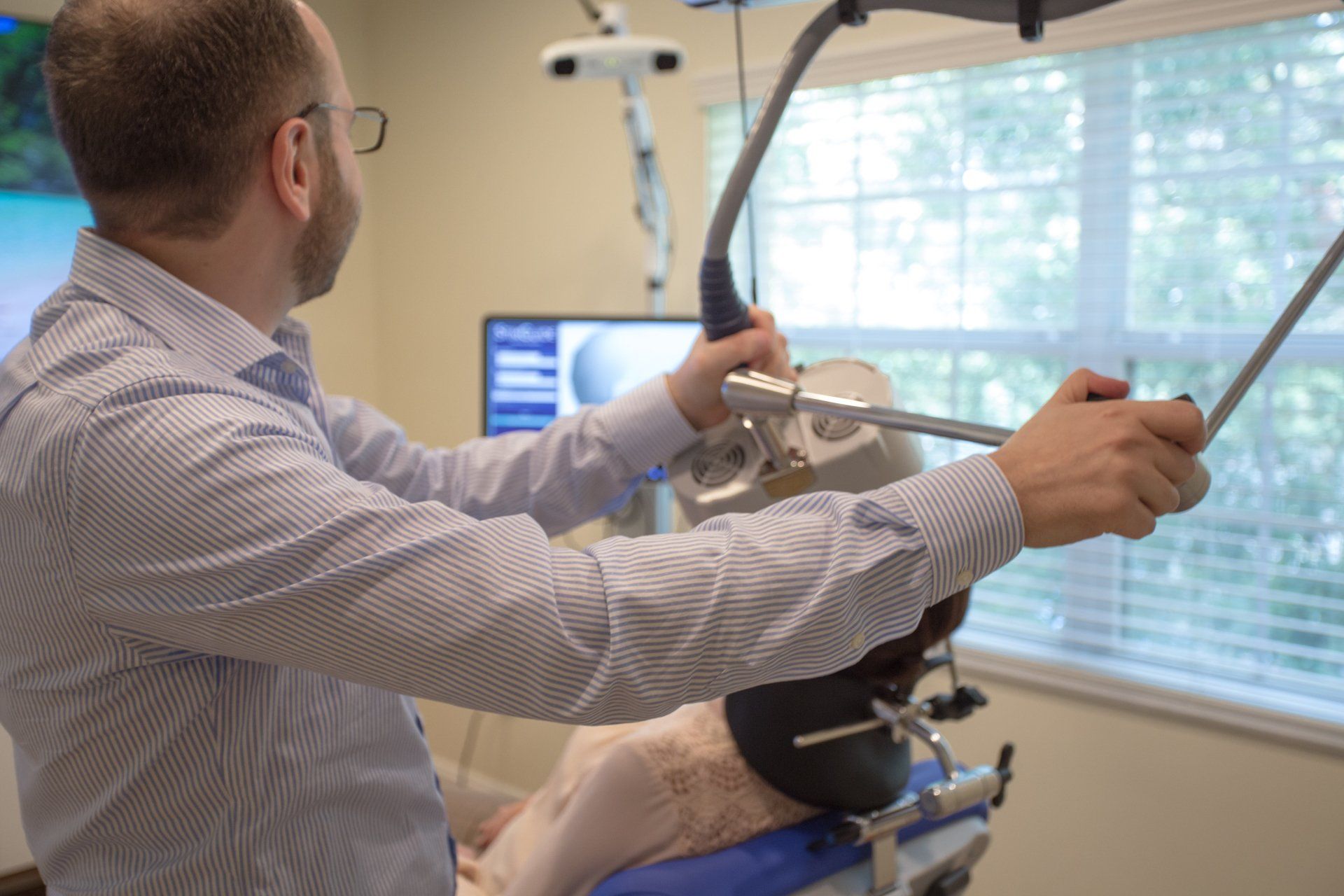 A doctor adjusting medical equipment on a patient's head, near a window. The doctor is wearing a striped shirt.